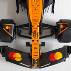 A top-down view of a McLaren Formula 1 car nose section, featuring a bright orange and black livery with sponsor logos including Hilton, Mastercard, and webex, set against a white textured background.
