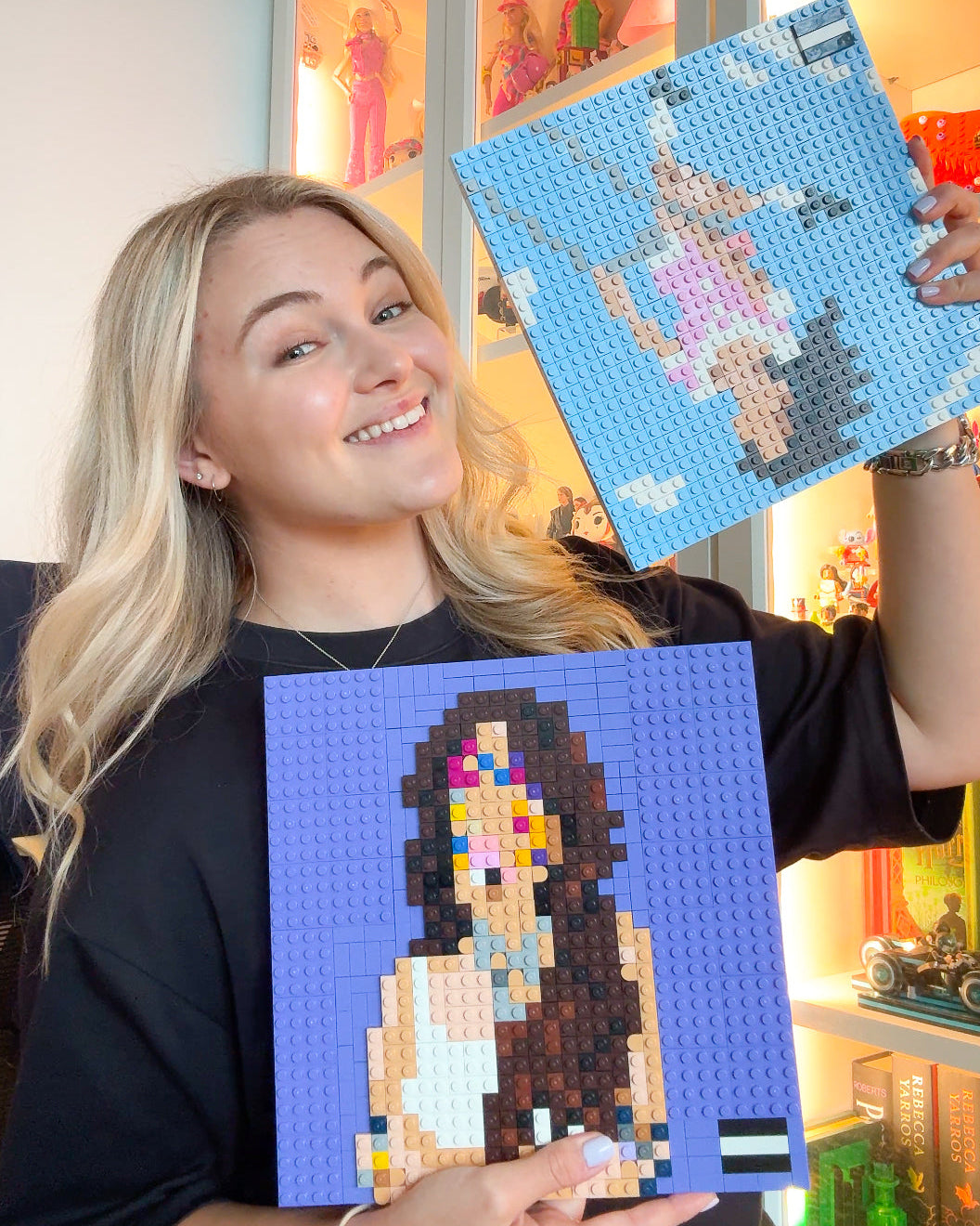 A smiling blonde woman holds up two custom pixel-art brick building toy portraits. The portrait on the left is on a blue background with dark hair, and the portrait on the right is on a light blue background with a pink dress.